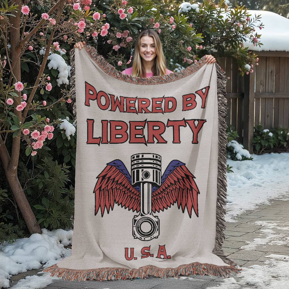 Customer holding up the Powered by Liberty Woven Blanket outdoors, showcasing the patriotic piston-and-wings design in red, white, and blue—crafted from 100% woven cotton and made in the USA