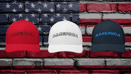 Three AMERICA arched trucker hats in red, white, and navy, featuring bold embroidered stitching in matching colors, displayed against a patriotic American flag brick wall background.