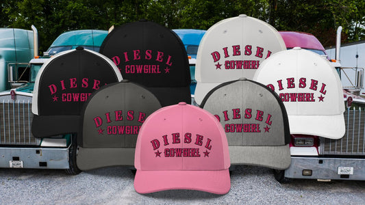 A collection of Diesel Cowgirl trucker hats in various colors, including black, white, pink, and grey, with bold pink embroidered lettering, displayed in front of a lineup of classic semi-trucks.