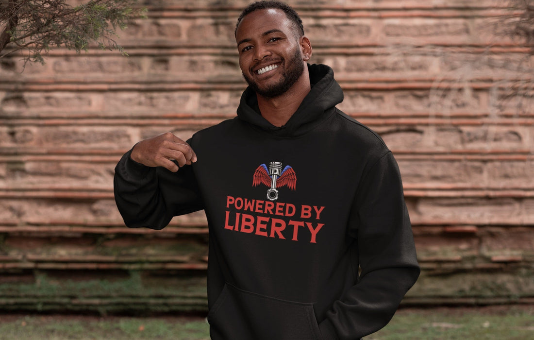 Man wearing a black 'Powered by Liberty' patriotic hoodie featuring an engine piston with red and blue wings. Rugged American flag-inspired design for truckers, bikers, and patriots.