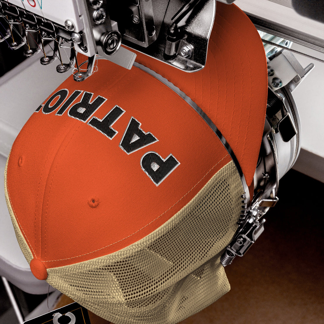 An orange and khaki mesh trucker hat being embroidered with the word 'PATRIOT' on a professional stitching machine, showcasing high-quality craftsmanship and attention to detail.