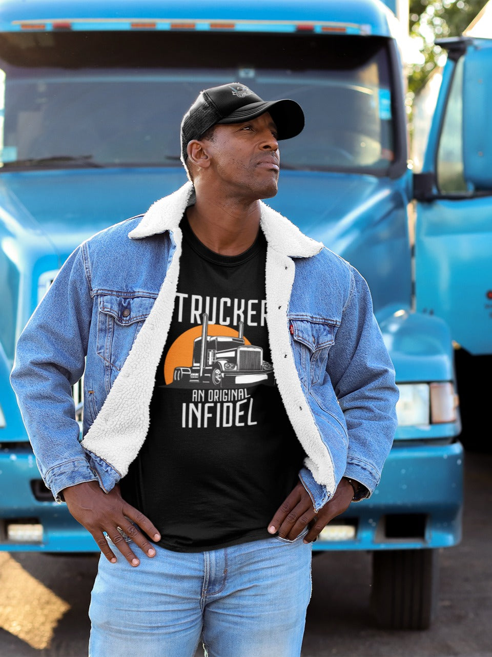 Man wearing a 'Trucker - An Original Infidel' graphic t-shirt, denim jacket, and trucker hat, standing confidently in front of a blue semi-truck.