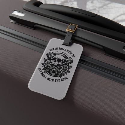 Luggage tag featuring a skull, pistons, and roses with the phrase "Death Rolls With Us – Get Right With the Road", attached to a suitcase.