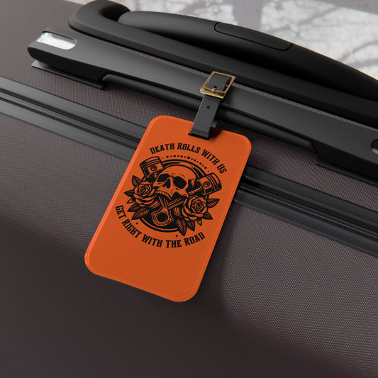 Luggage tag featuring a skull, pistons, and roses with the phrase "Death Rolls With Us – Get Right With the Road", attached to a suitcase.