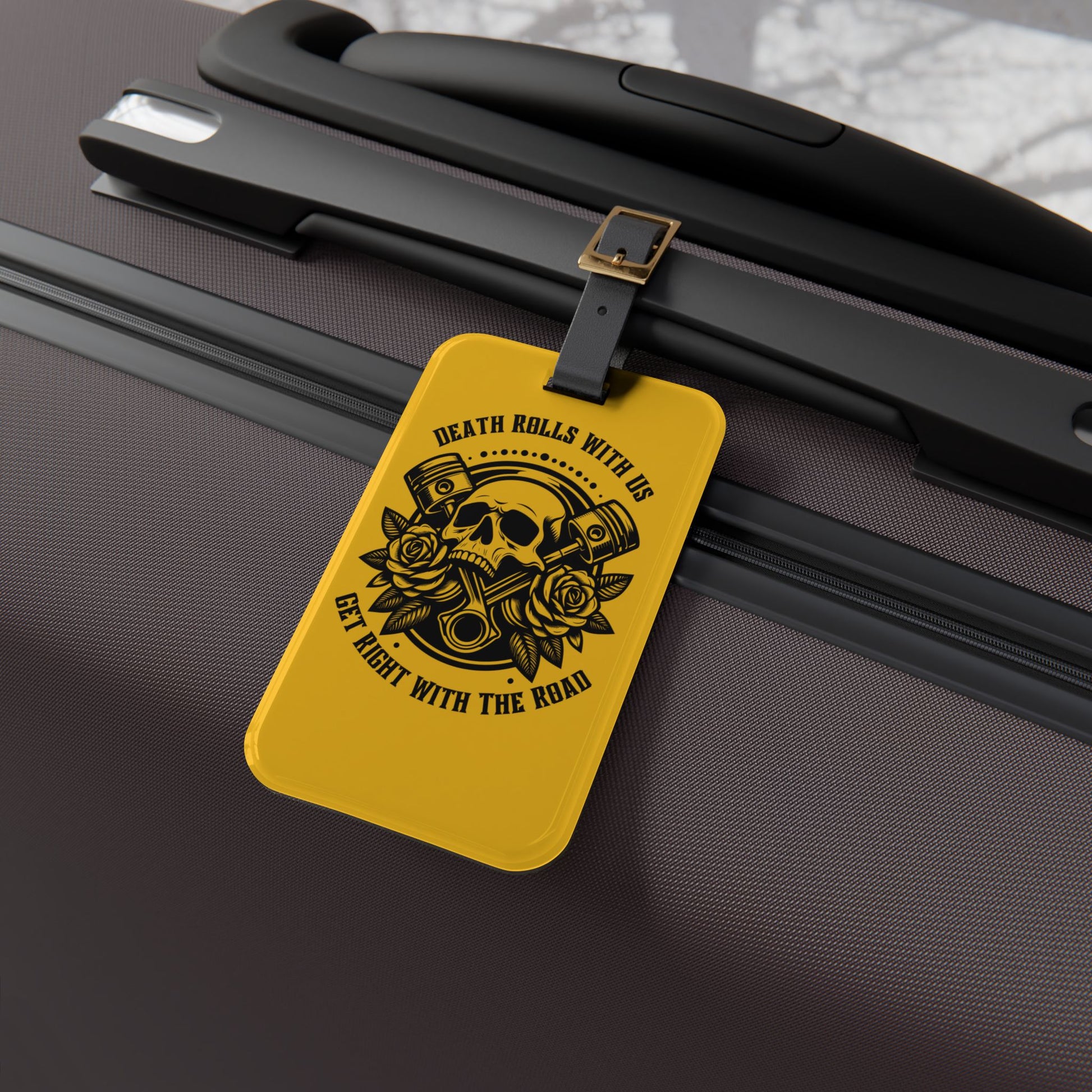 Luggage tag featuring a skull, pistons, and roses with the phrase "Death Rolls With Us – Get Right With the Road", attached to a suitcase.