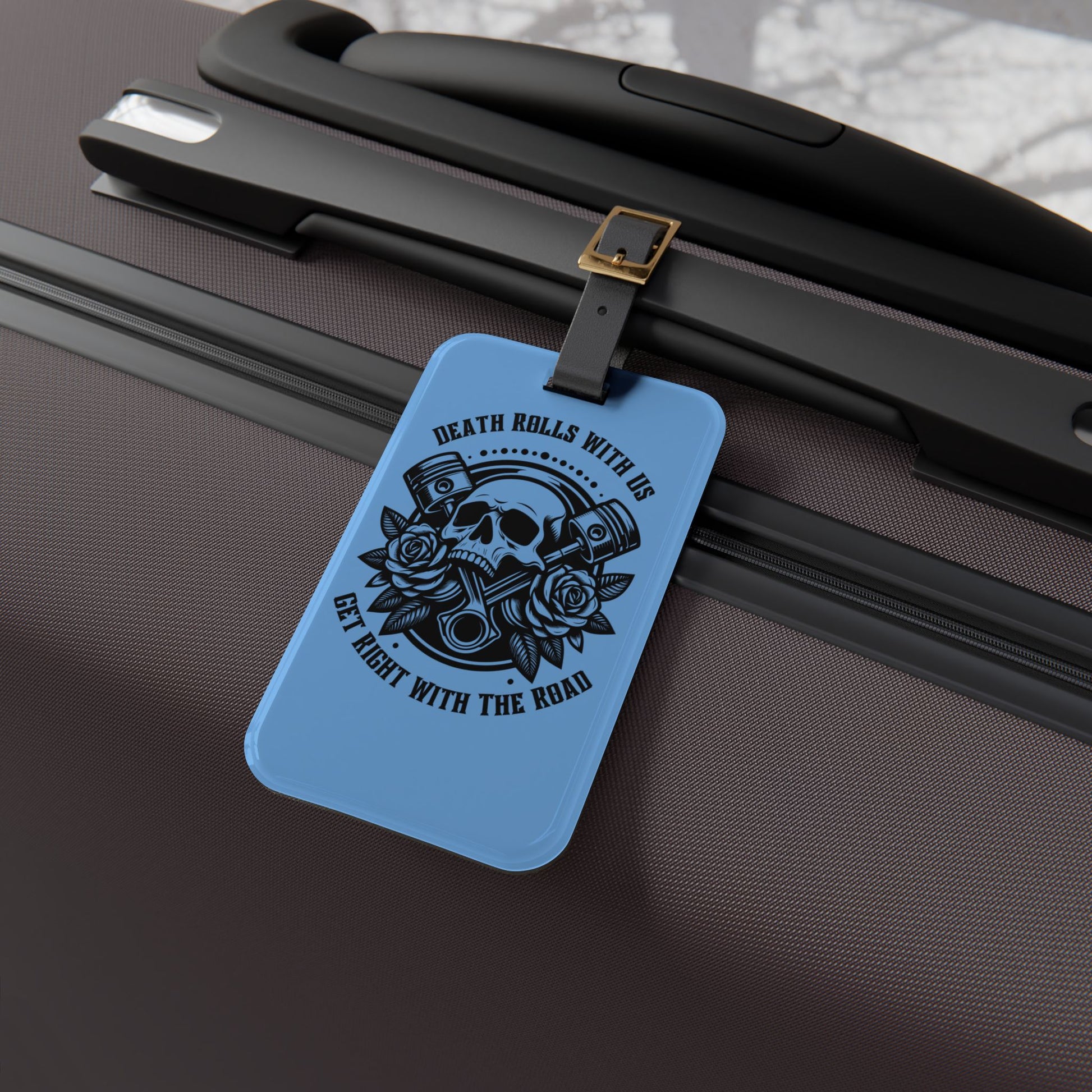 Luggage tag featuring a skull, pistons, and roses with the phrase "Death Rolls With Us – Get Right With the Road", attached to a suitcase.